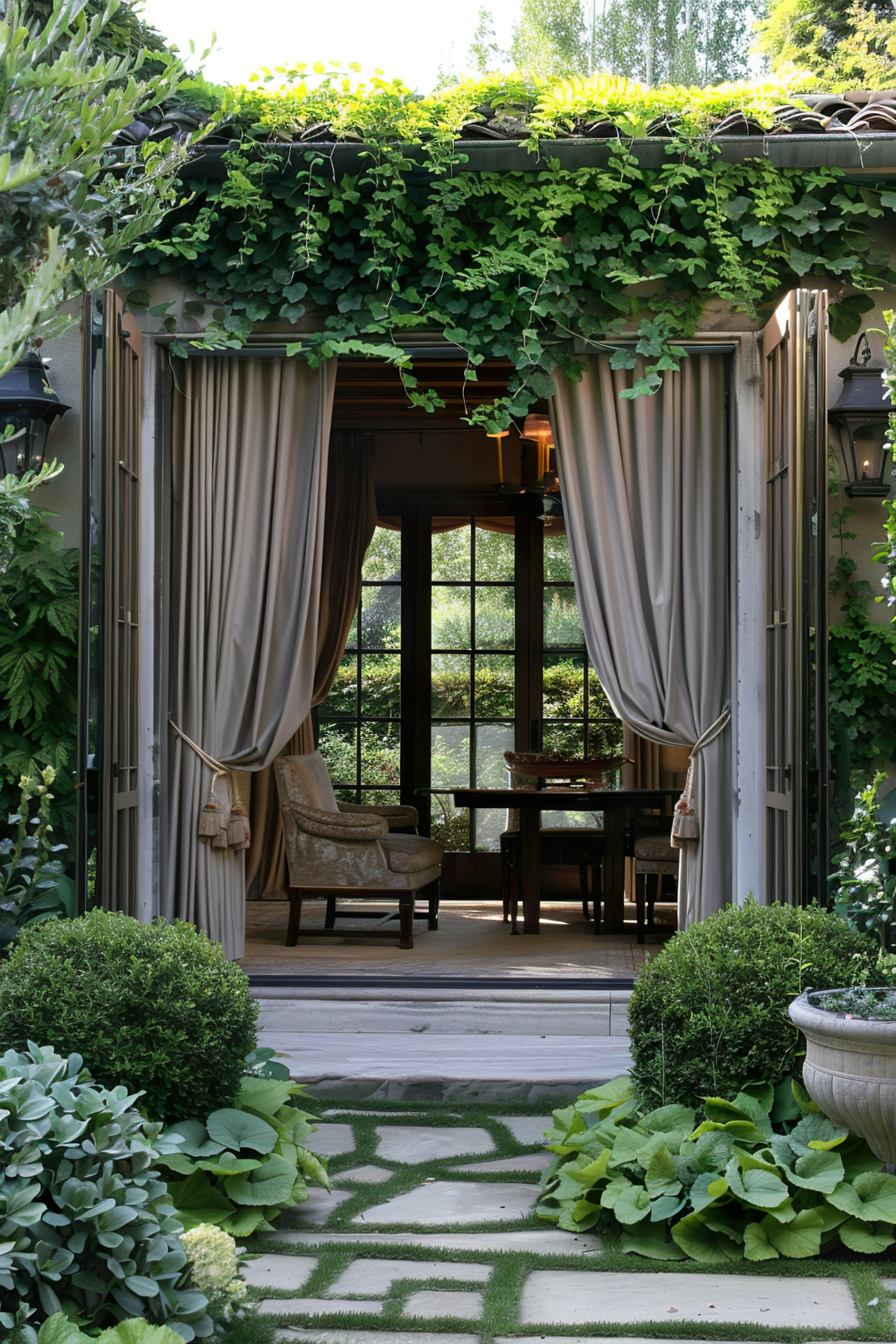 A serene garden patio with draped curtains leading to a cozy room, surrounded by lush greenery and patterned stone pathway.