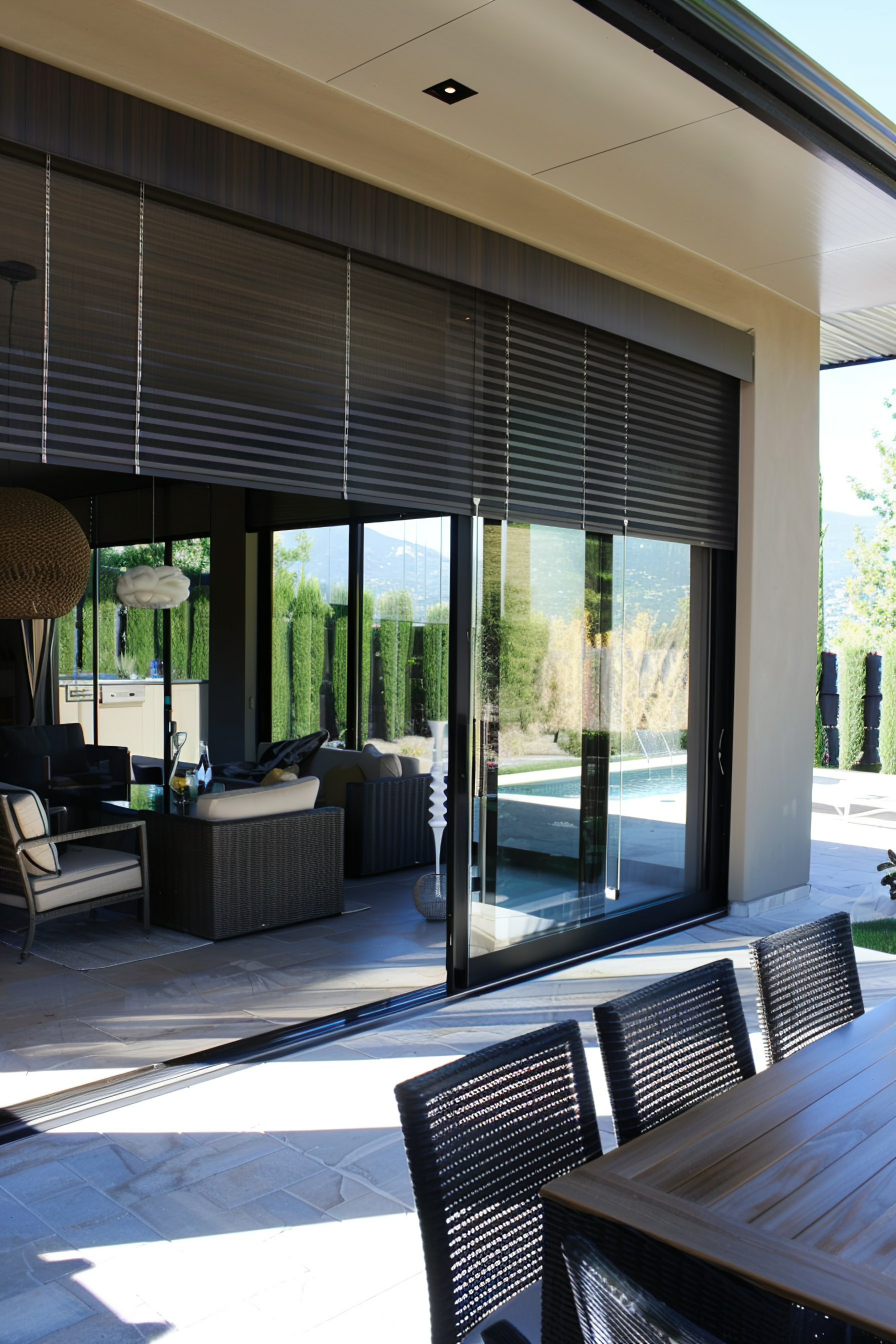 Modern outdoor patio with furniture, leading to an interior living space through large glass doors, overlooking a swimming pool.