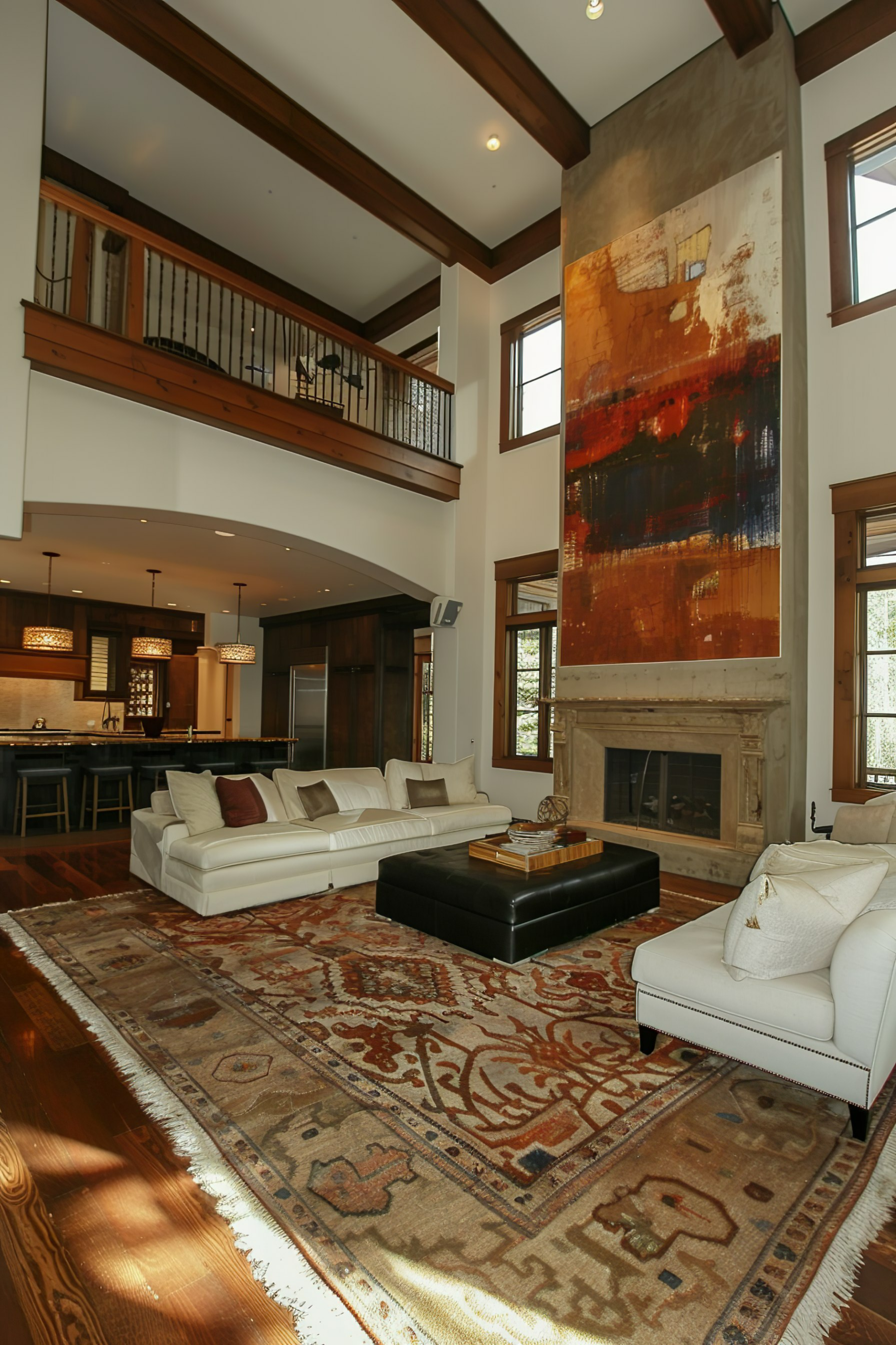 Interior of a spacious living room with high ceilings, large abstract painting, white sofas, an ornate rug, and a kitchen in the background.