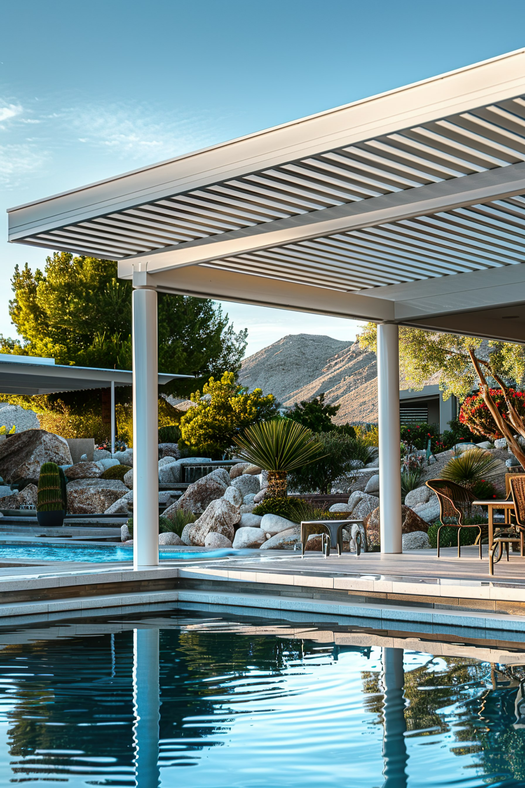 An elegant poolside with a modern pergola, outdoor furniture, and a landscaped rocky backdrop under a clear sky.