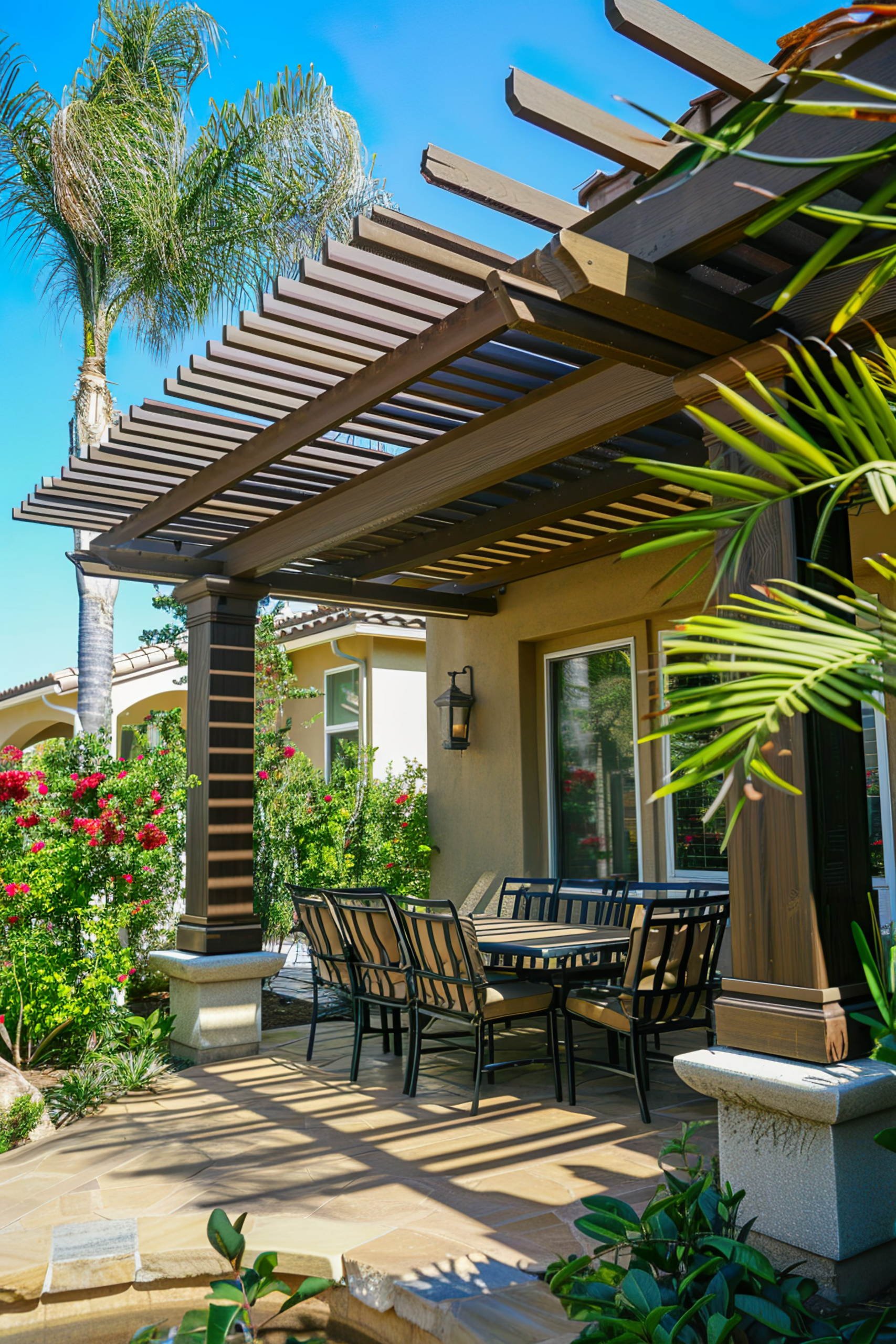 A sunny patio with a pergola, outdoor dining set, blooming bushes, and a palm tree evoking a tranquil backyard setting.