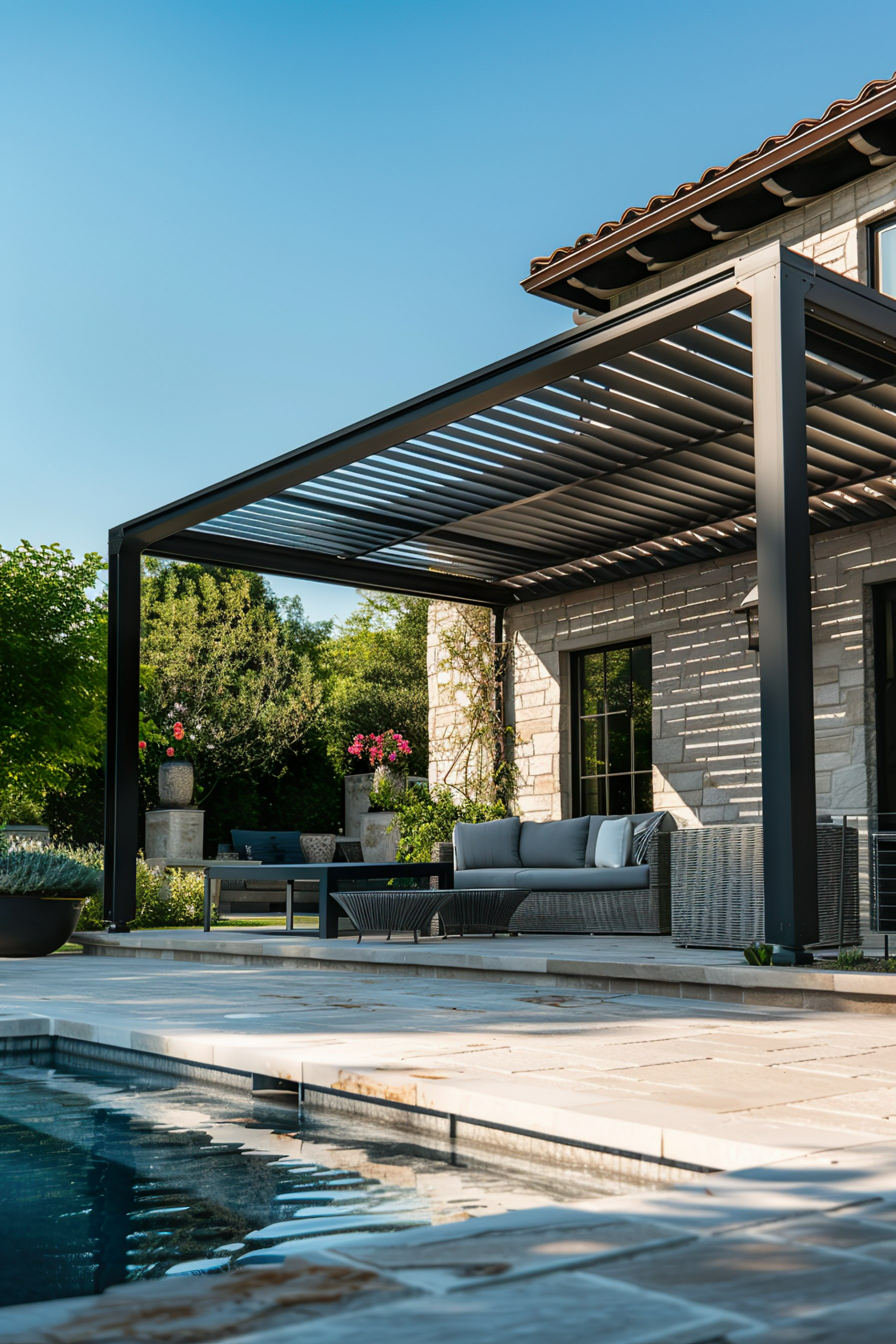 Luxurious backyard patio with modern furniture under a pergola, next to a swimming pool, and surrounded by greenery.