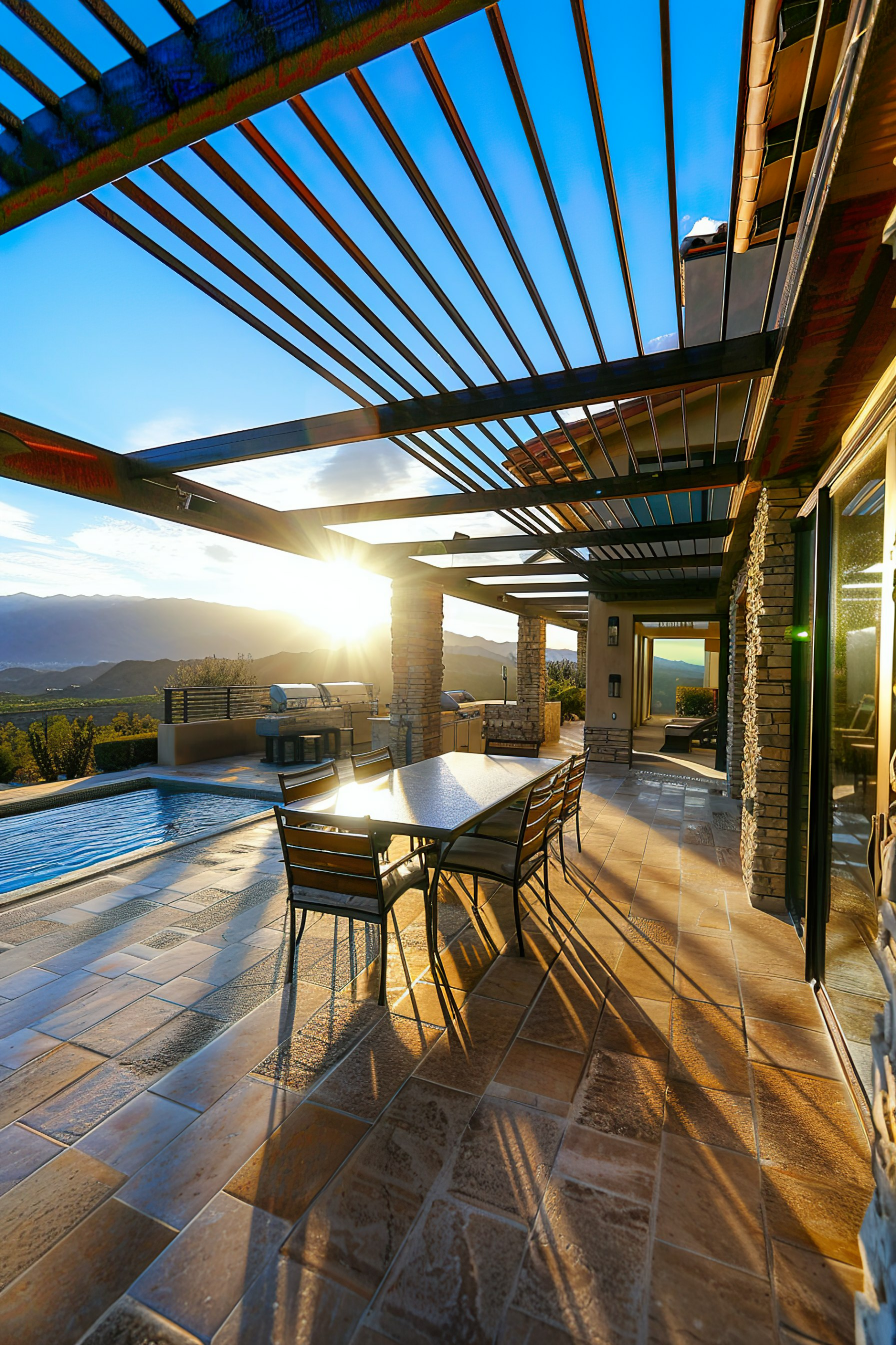 Luxurious patio with dining area overlooking a pool and mountains at sunset, with sunlight casting dramatic shadows.