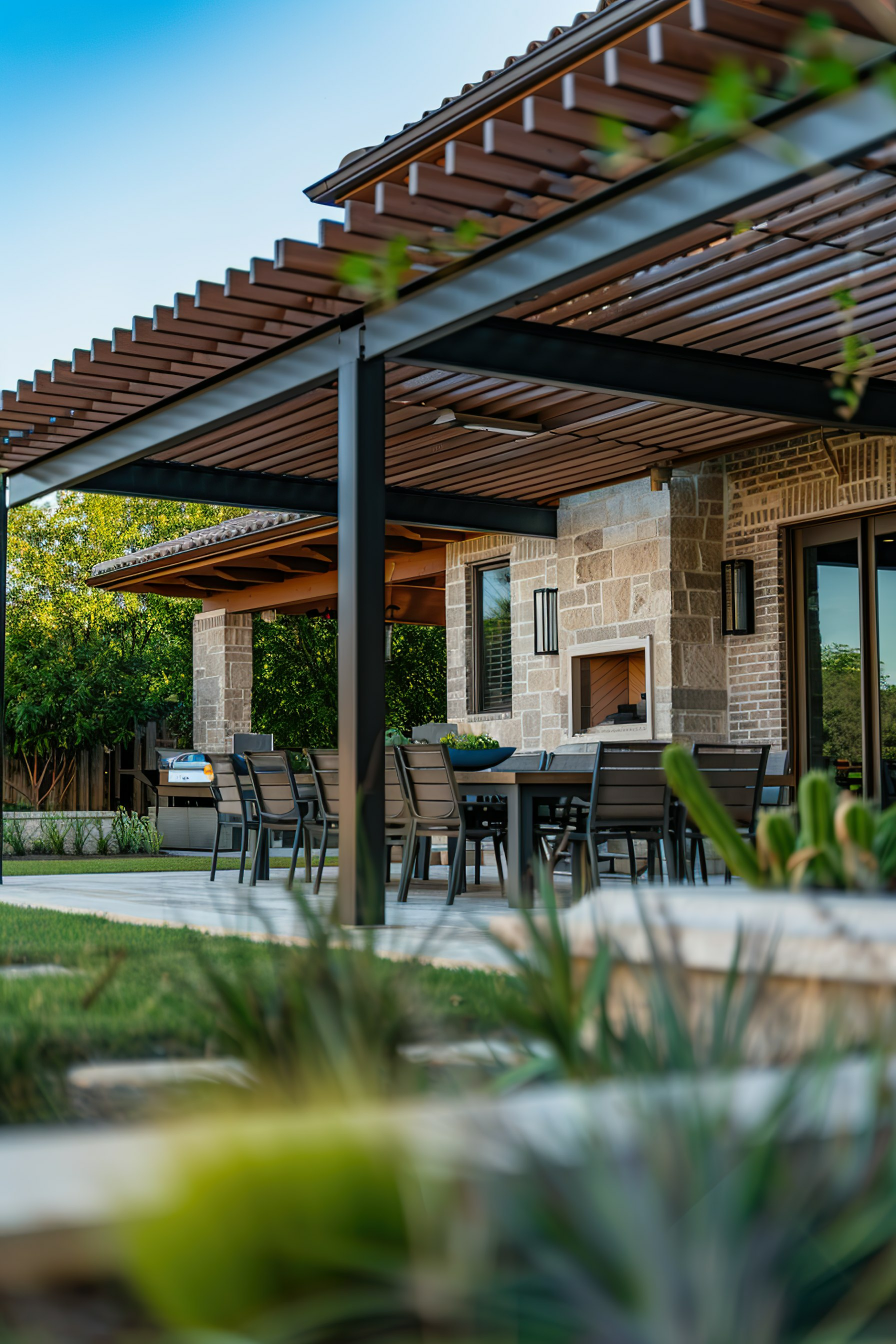 ALT: Elegant outdoor patio area with dining table and chairs under a modern pergola, adjacent to a stone-faced house.