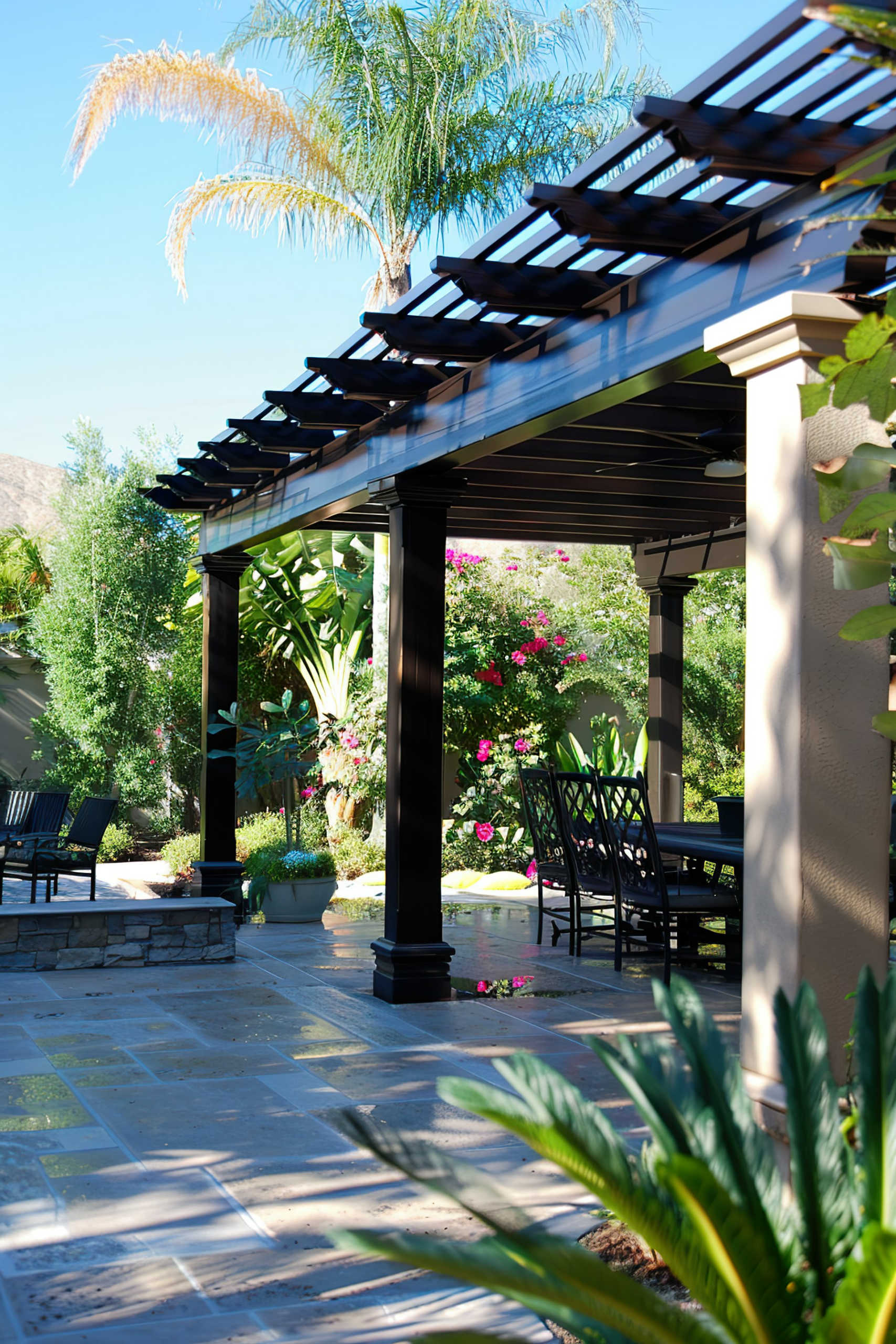 A tranquil patio area with a pergola, stone flooring, lush greenery, and outdoor furniture under bright blue skies.