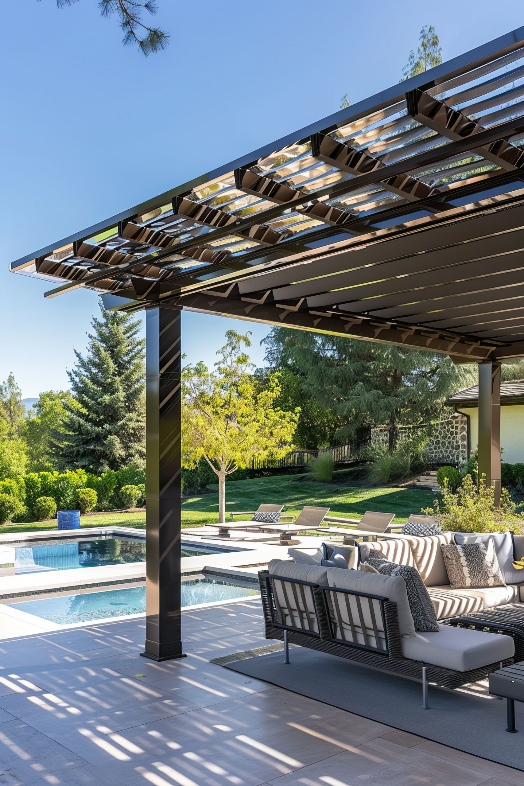 Outdoor patio with modern furniture under a pergola, beside an in-ground pool, with lush trees in the background.